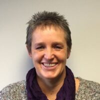 A person with short hair smiling, wearing a gray sweater and a purple scarf, stands against a plain light-colored background.