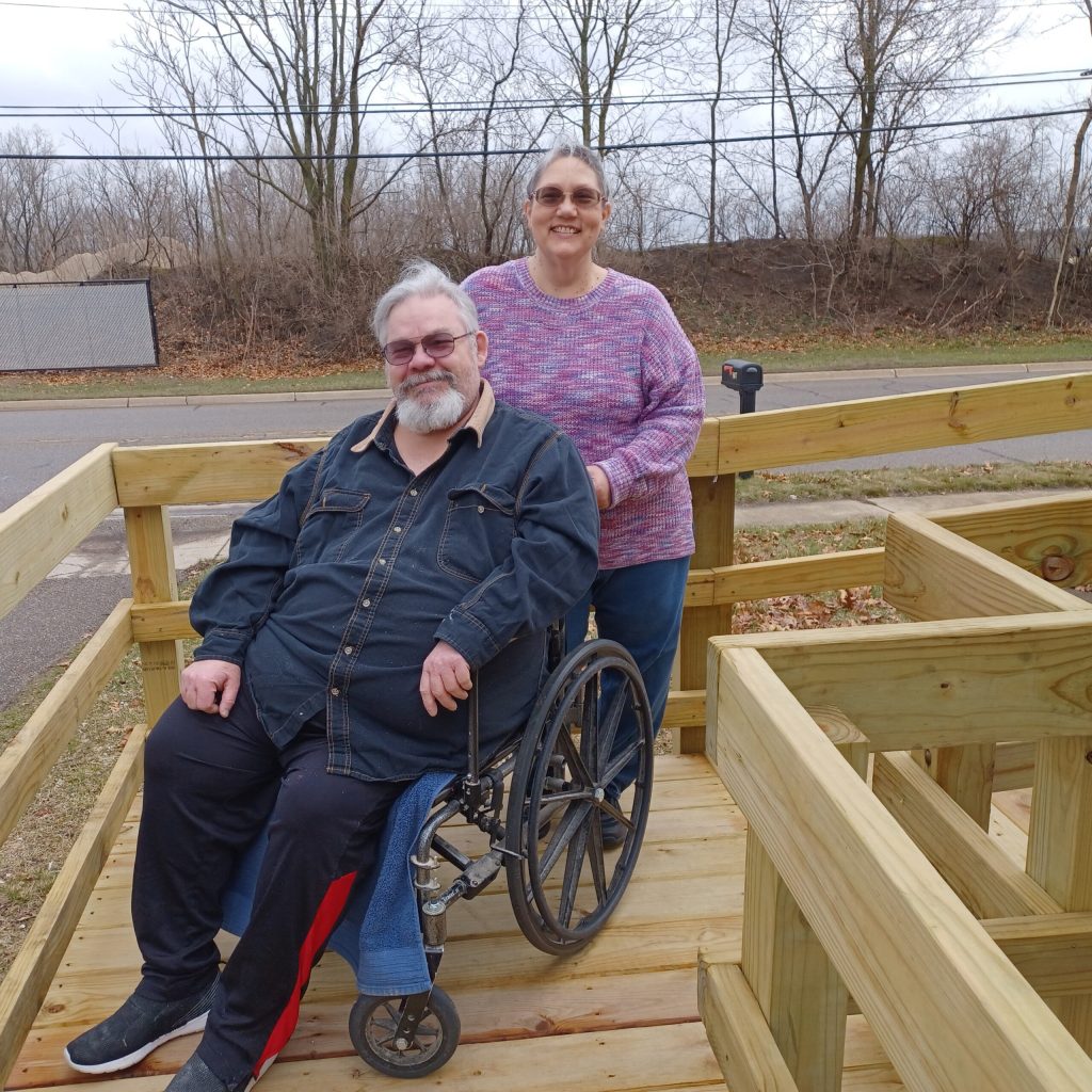 A person is sitting in a wheelchair on a wooden ramp, and another person is standing behind them.