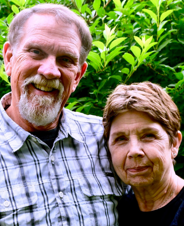 An older couple stands close together smiling in front of green foliage. The man wears a plaid shirt, and the woman has short hair and a dark top.