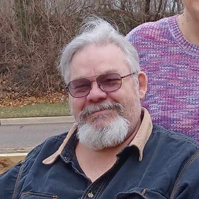 Smiling older man with sunglasses and a beard, outdoors, next to a person in a purple sweater.