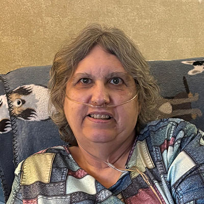 A woman with an oxygen tube smiling while sitting on a patterned blanket-covered chair.