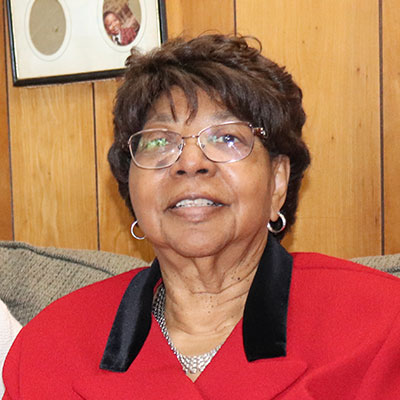 An older woman wearing glasses and a red jacket sits on a couch, with wood-paneled walls in the background.