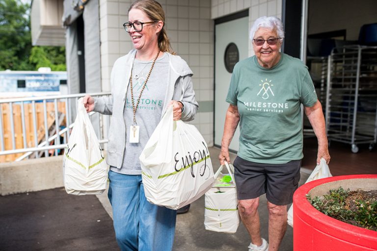 2 women wearing Milestone shirts, carrying bags of food