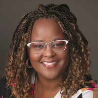 Smiling person with glasses and braided hair wearing a colorful top against a neutral background.