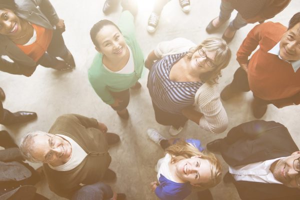 A diverse group of people standing in a circle, looking up with smiles, in a bright, sunlit space.