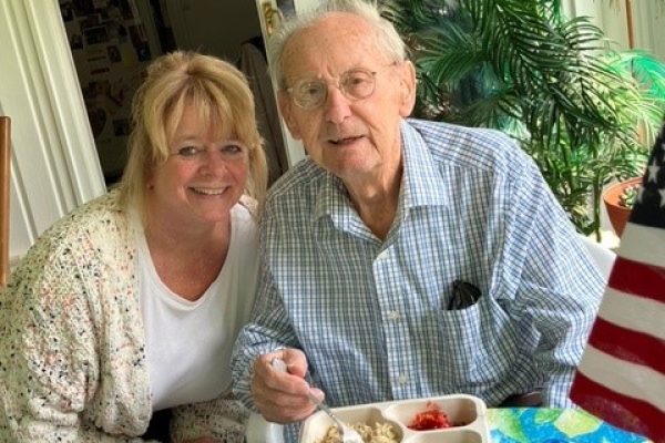 Smiling elderly man eating at a table with a woman beside him, an American flag and plants in the background.