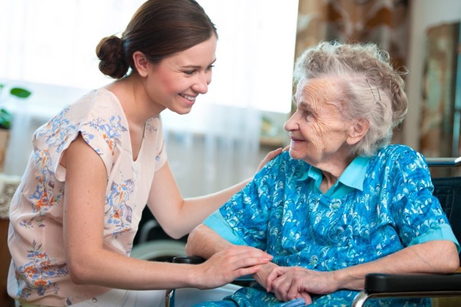 Young woman smiling and holding hands with an elderly woman sitting in a wheelchair indoors.