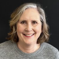 A woman with shoulder-length hair smiles at the camera against a black background, wearing a gray sweater.